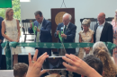 Two men in suits cut a green ribbon while several others look on