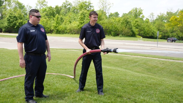 a man holds a fire hose while a more experienced firefighter watches