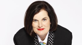 A woman with dark hair leans over a stool wearing a black blazer, black pants, white button-down shirt and blue patterned tie.