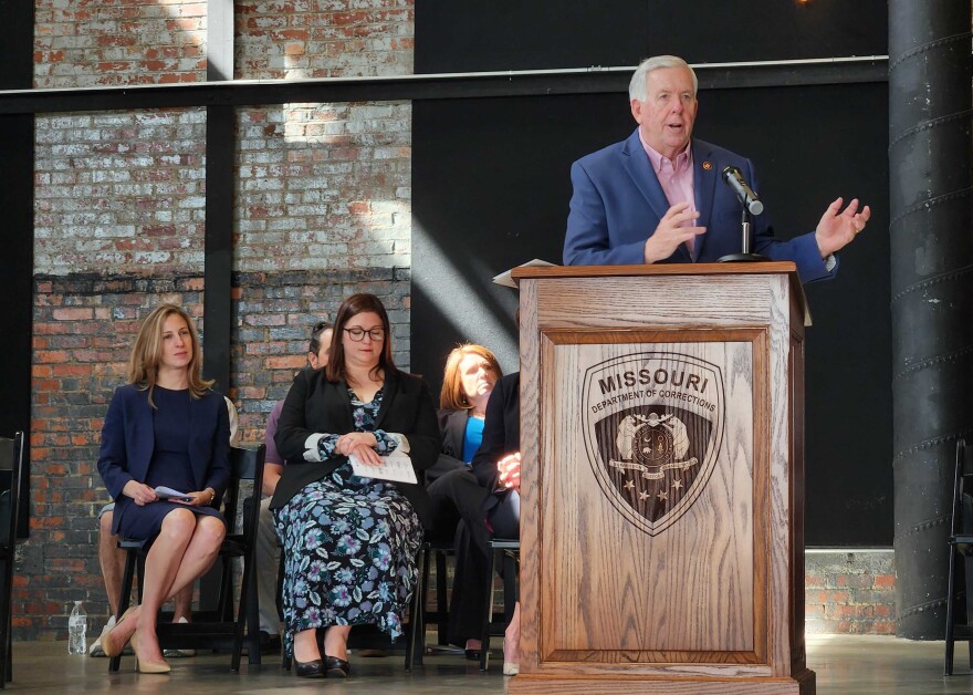 Gov. Mike Parson stands at a podium with a microphone on it. People sit behind him and look at him while he speaks.