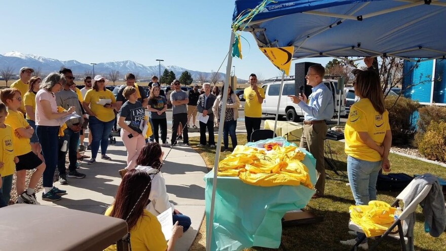 A man stands under a tent talking to people who are wearing yellow shirts. 