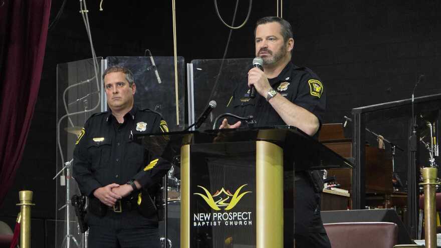 Two men in police uniforms. One is at a podium holding a microphone