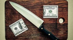 A hundred dollar bill cut in half with a knife on a cutting board.