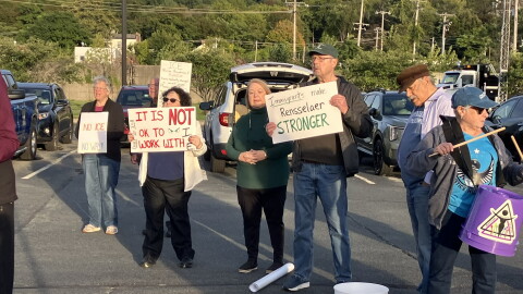 Several protesters are standing in a row in a parking lot. Most of them are holding posters, some of which say "It is NOT ok to with ICE" and "Immigrants make Rensselaer STRONGER." One woman is holding drum sticks and has a plastic bucket hanging from a strap around her neck. 