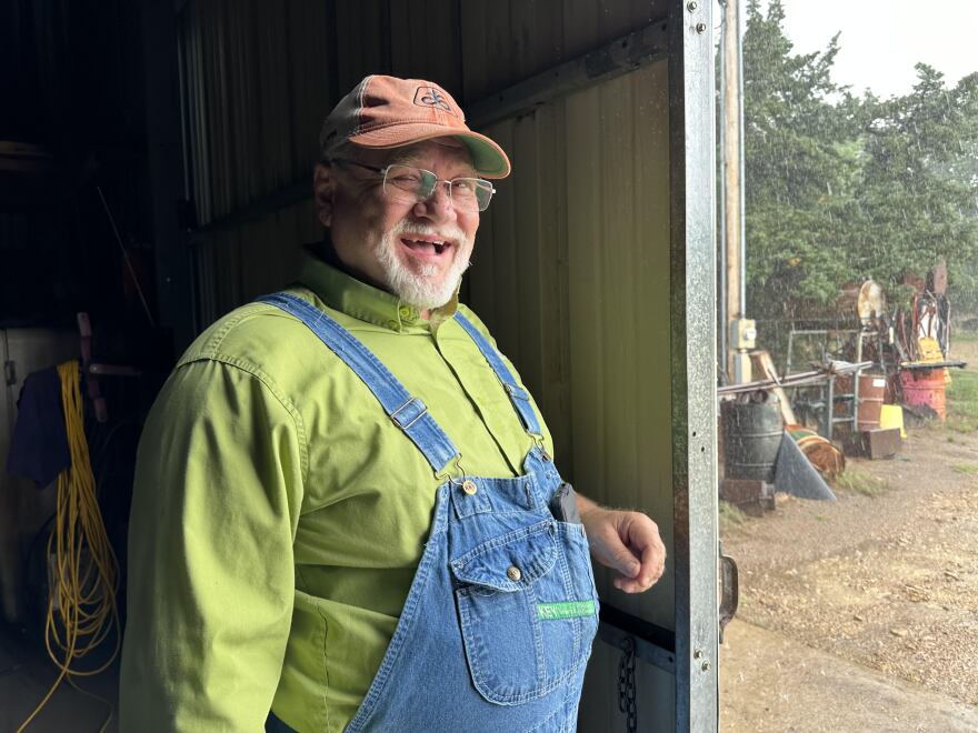 Kansas Farmer stands inside a shed, smiling as hard rain falls outside.