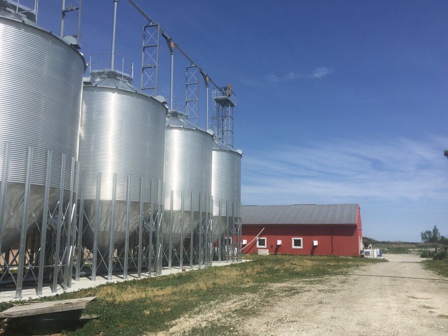 Silver silos sit in a row, leading up to a massive red dairy barn, with a dirt farm road between them. The sky is blue and it is early summer.