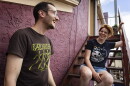 Two people who look to be in their 30s laugh while standing and sitting on the stairs outside a burgundy building.