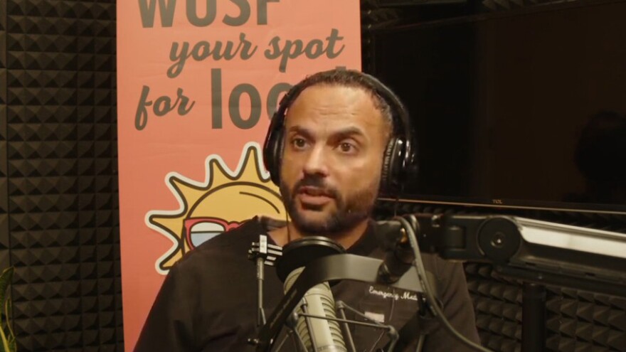 Man with short dark hair and beard speaking into a microphone while sitting