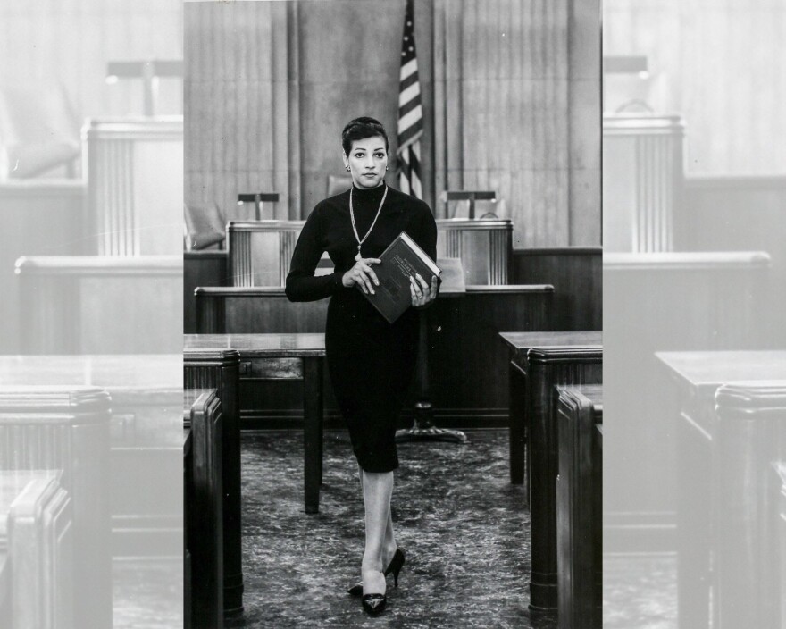 A young Judge Lucile A. Watts in a black dress stands confidently in a courtroom. She holds a book, with an American flag and judge's bench in the background. The image is in black and white.