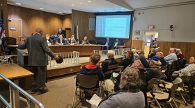 A man stands facing away from the cap, holding a cap with arms spread wide, addressing a panel of five local government officials in front of him, and fellow residents behind him.