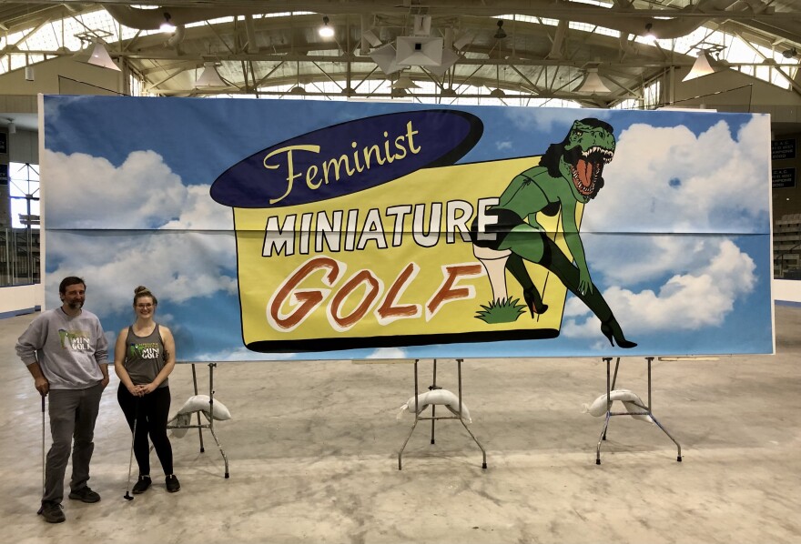  Colin Boyd and Carly Thomsen standing in front of a large sign saying, "Feminist Miniature golf."