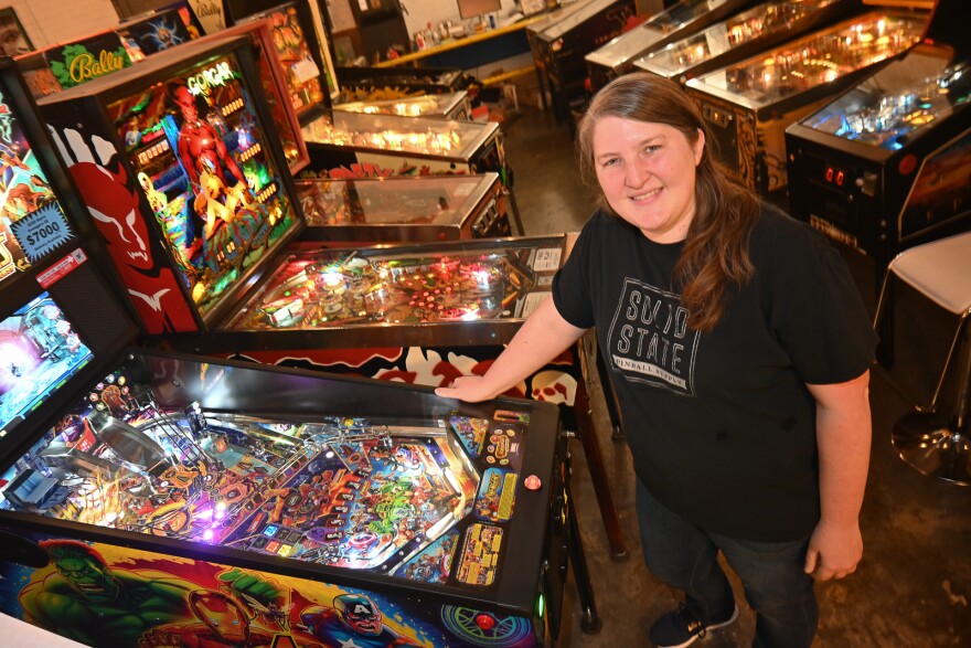 A woman stands next to a pinball machine. She is looking at the camera. Behind her are many pinball machines lined up in two rows.