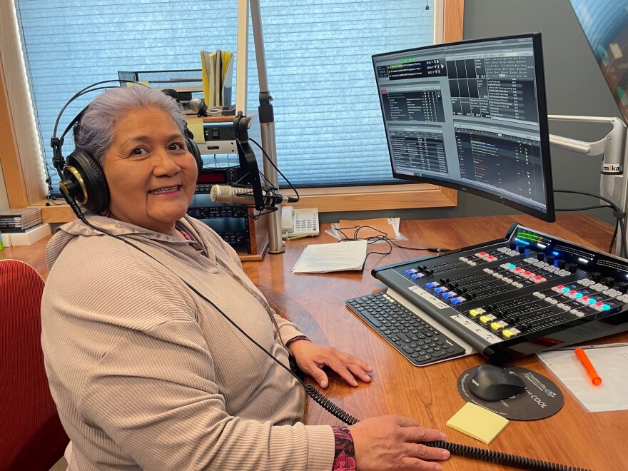 A woman sits in front of a microphone, smiling at the camera. A sound board and computer monitor are in front of her.