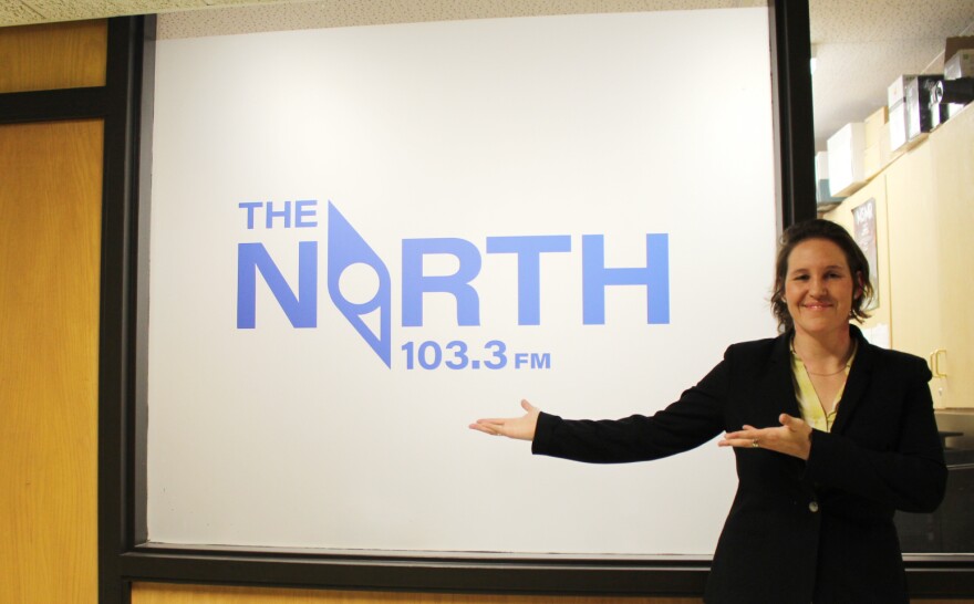 Woman standing in a hallway to the right of a frosted window that says The North 103.3 FM in blue lettering, gesturing to the lettering with both arms and smiling.