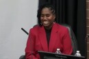 Woman seated at table with microphone and wearing a red jacket and black blouse