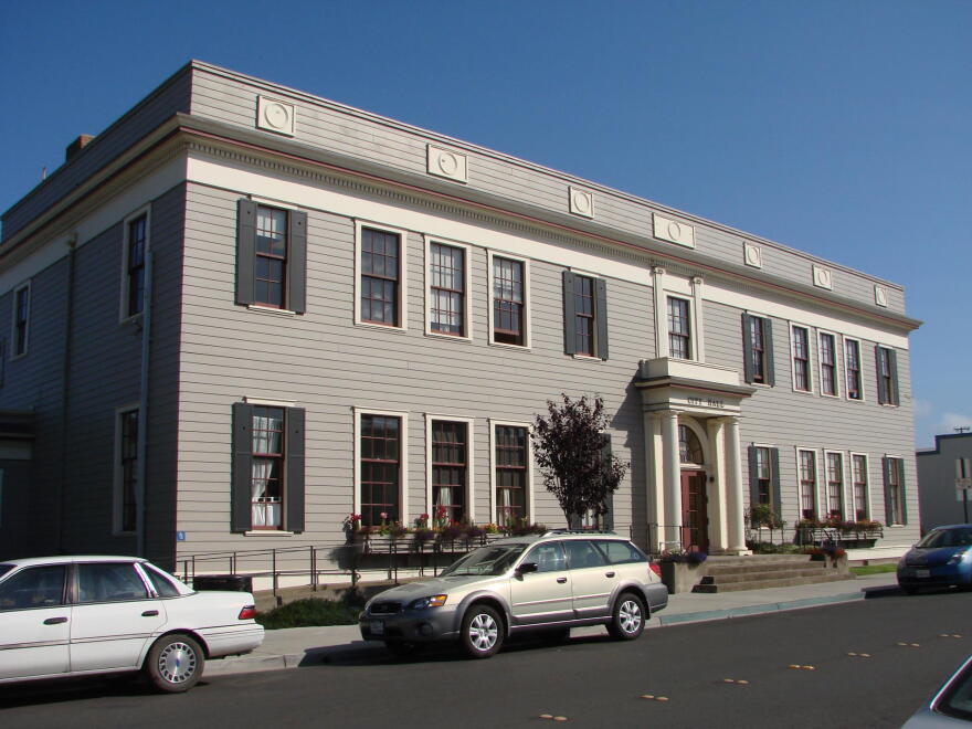 A gray two-story building with a rounded red doorway, seen from across the street with cars in front.