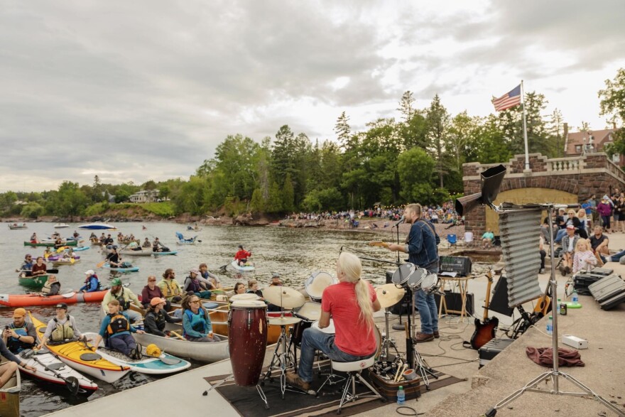 Two men performing on a pier to a crowd of people on the shore and people in kayaks and canoes. 