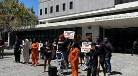 Several young people standing in a semi-circle in front of the administration building. Two are in orange jumpsuits and one individual is on a step ladder with a bull horn.