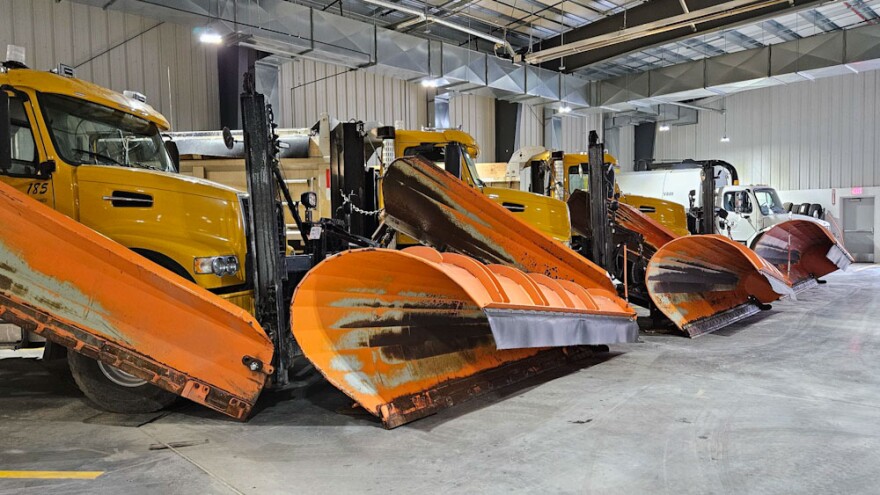 A dump truck sits in the foreground of a large garage, with a yellow cab that has two orange snowplow attachments, while several identical trucks recede into the background.