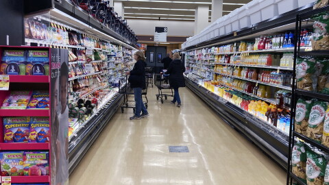 Shoppers with carts stand in a grocery store aisle, surrounded by products on both sides.