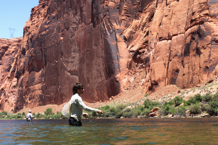 A man stands thigh-deep in a river casting a fishing line. Behind him are steep, reddish rock formations.