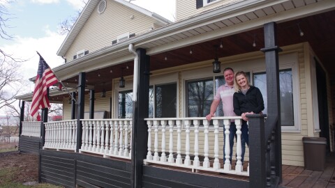 Elizabeth and Chad Pierce stand on their porch, an American flag hangs off the porch at the far left of the image. 