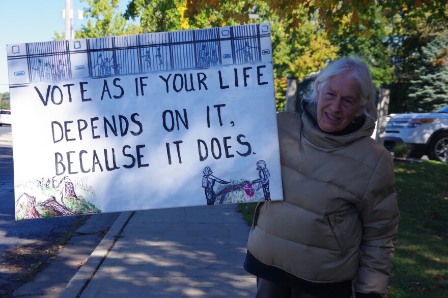 A woman in a tan puffer jacket on Drake Road stands in the shade of a tree holding a white sign that reads in black letters "Vote as if your life depends upon it because it does." 