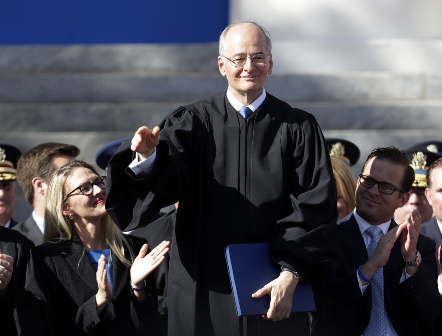 A man in a black judge's robe waves as onlookers clap