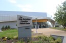 Exterior of the Doug Oberhelman Caterpillar Visitors Center, a modern building with angular, sail-like metal paneling and a large monument sign reading "The Doug Oberhelman Caterpillar Visitors Center" alongside the Caterpillar logo. Glass doors and windows with photo displays are visible under a bright yellow awning band.