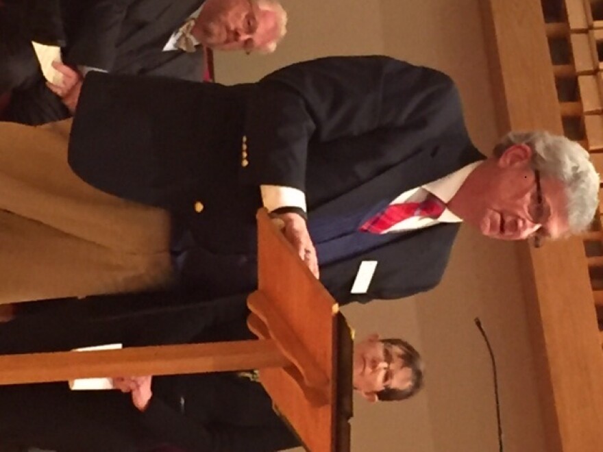 Man at a podium with another man to left and a woman to his right.