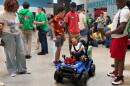 A woman stands looking at a child seated in a battery-operated toy car. The child is looking up and smiling at a man who is looking down and smiling at the child. Other people are standing an sitting in the background