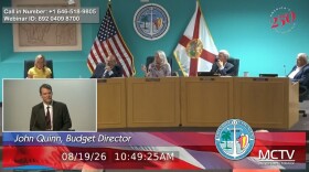 John Quinn appears in a tiny window on the lower-left of the screen with Monroe County Commissioners appearing on the dais behind him.