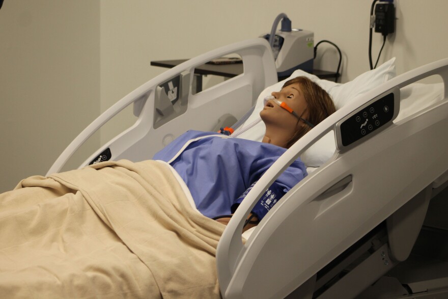 A mannequin used for medical training lays in a hospital bed. The mannequin wears a nasal cannula and a hospital gown with a blanket up to its waist. 