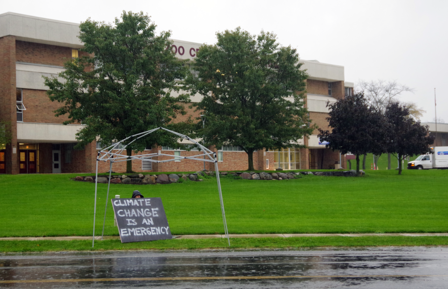 A man in a hooded raincoat sits behind a large sign that says "climate change is an emergency." He is sitting under a metal awning frame. Kalamazoo Central High School is in the background.