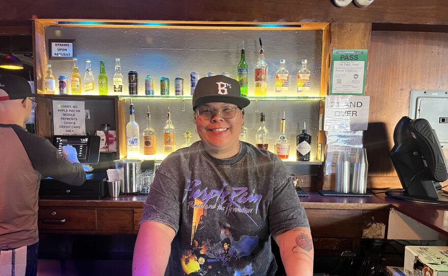 Patty stands behind the bar at the White Horse wearing an Oakland Ballers cap and a Prince t-shirt, smiling.