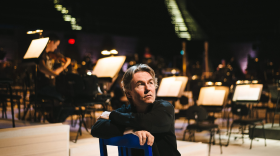 Esa-Pekka Salonen sits on stage before a concert and looks off into the distance. Behind him, stage lights lend a glow to the stage.