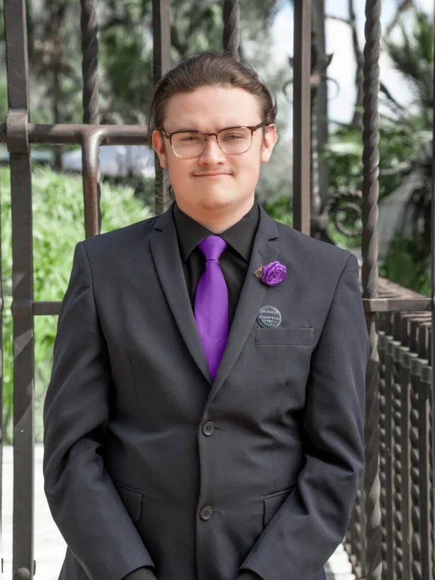 Young person in black suit with purple tie wearing glasses. 
