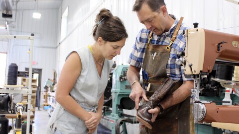 A man in a buttoned-down shirt and apron holds up a cowboy boot being stitched together to show a woman in a denim jumper.