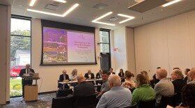 An audience sits in front of a panel. There is a projection screen overhead