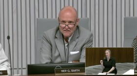 A middle aged bald man wearing a gray suit jacket and glasses sits at a dais and speaks into a microphone. 