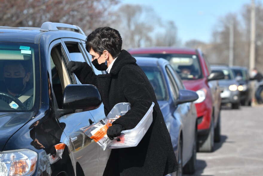 A woman in a black coat carries a plastic bin filled with plastic bags. She is reaching into the driver's side of a car.  A line of cars sits behind her while another person in the background reaches into a car.