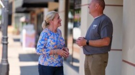 A woman and man stand on a sidewalk smiling towards each other. 