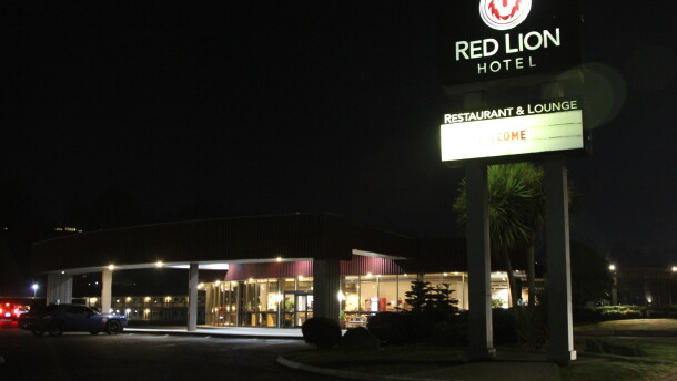 The lights through a hotel lobby window illuminate a dark parking lot at nighttime and a large sign with the words, "Red Lion Hotel." 