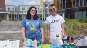 Two people stand behind a table with various items including a t-shirt, scrapbook open to newspaper articles, a plush smiling Earth toy and plastic bottles stuffed with more plastic material. The people are facing the camera and smiling