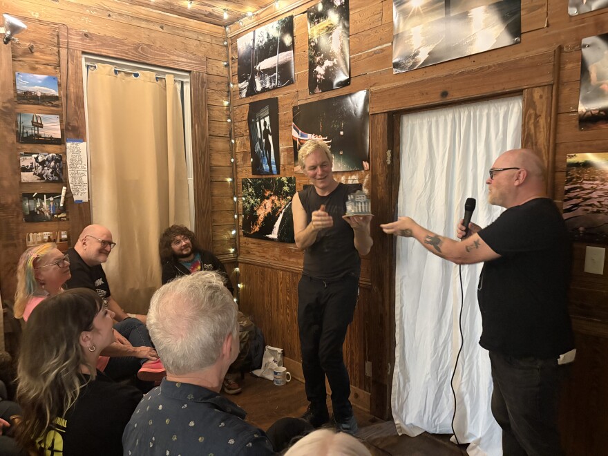Scott Satterwhite and Aaron Elliot (aka Aaron Cometbus) speak to a seated audience inside the 309 Punk Project house in Pensacola, as Cometbus holds a small model of the home created by an artist in residence.