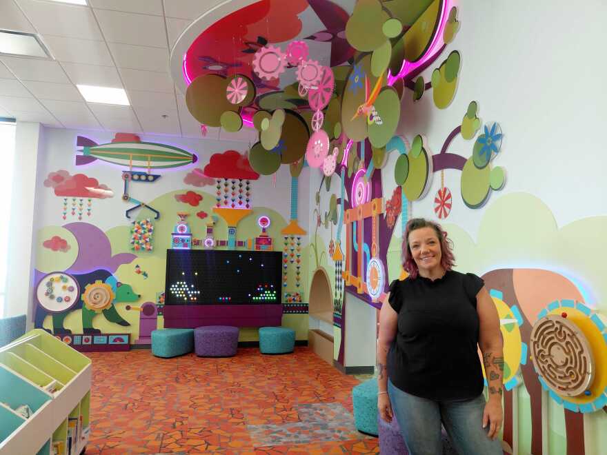 A women with purple hair, a black blouse and blue jeans stands in front a sensory friendly library space. 
