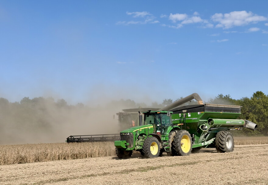 A green John Deere tractor pulling a wagon drives next to a combine harvesting soybeans.