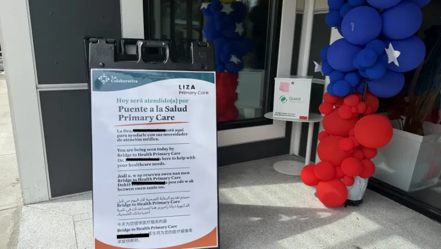 A sidewalk sign shows health information in multiple languages near some red and blue balloons.