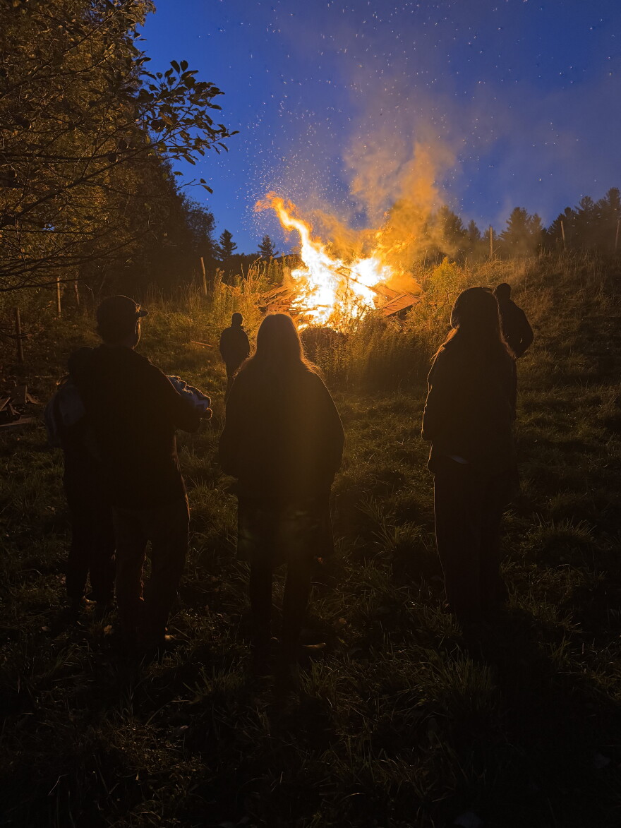 A bonfire against a blue sky with dark figures standing nearby