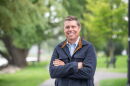 A smiling man with short brown hair standing with his arms crossed. He is wearing a blue collared button-down shirt under a blue fleece with a brown lining. 